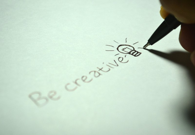 The words "Be Creative" being written onto a sheet of paper with a lightbulb doodle